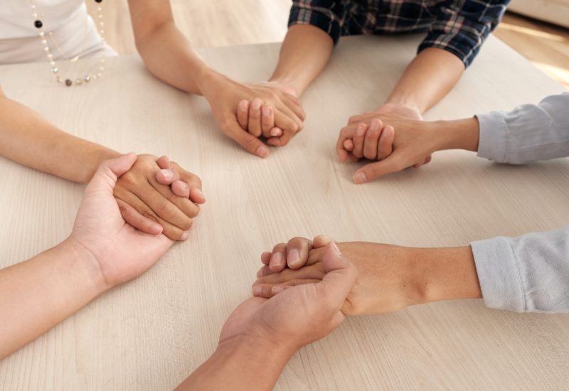 four-unrecognizable-people-sitting-around-table-holding-each-other-s-hands-middle